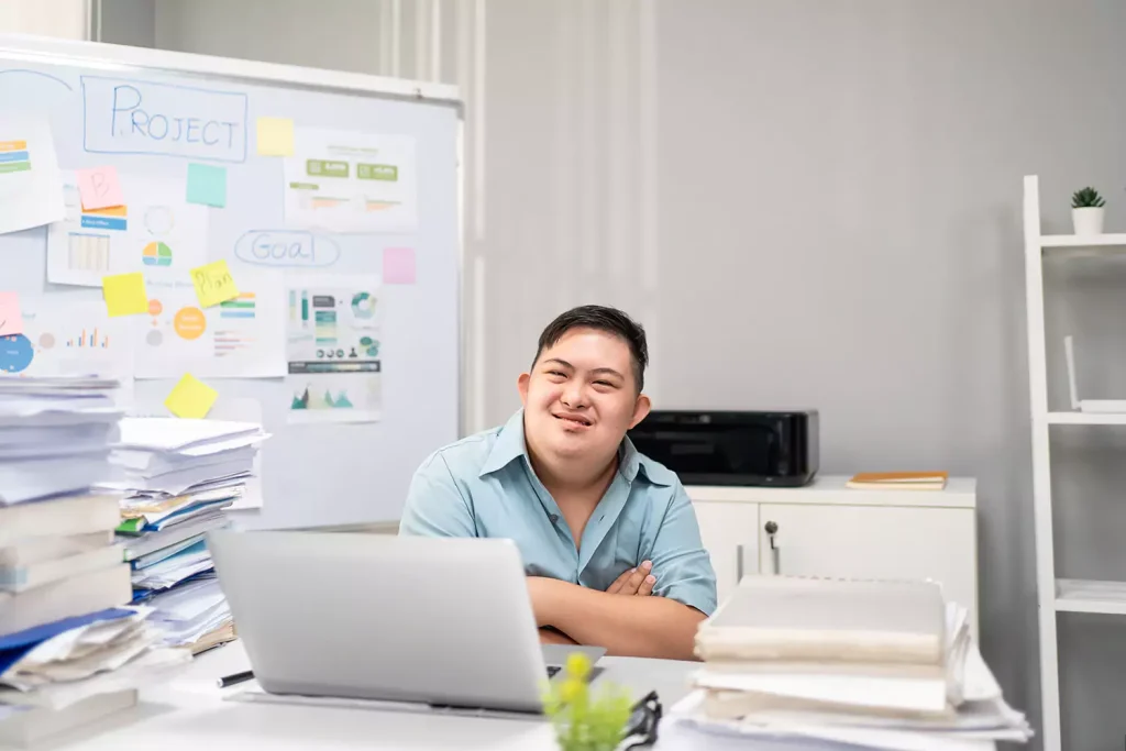 Disabled man with down syndrome sitting in office with laptop and smiling at camera
