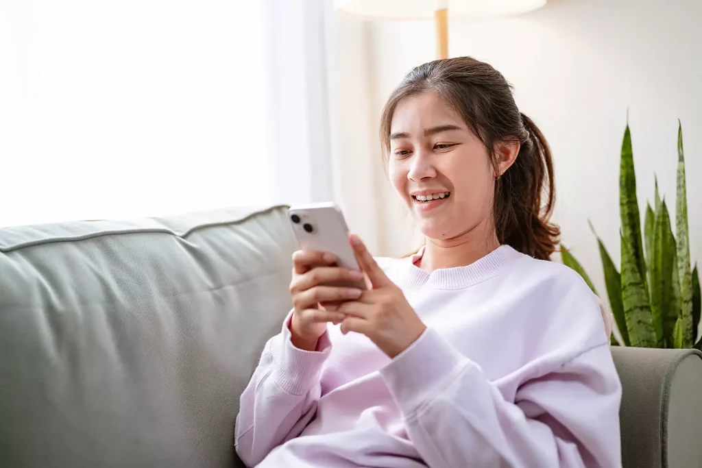 Happy Asian woman sitting on couch using mobile phone to look up accommodation